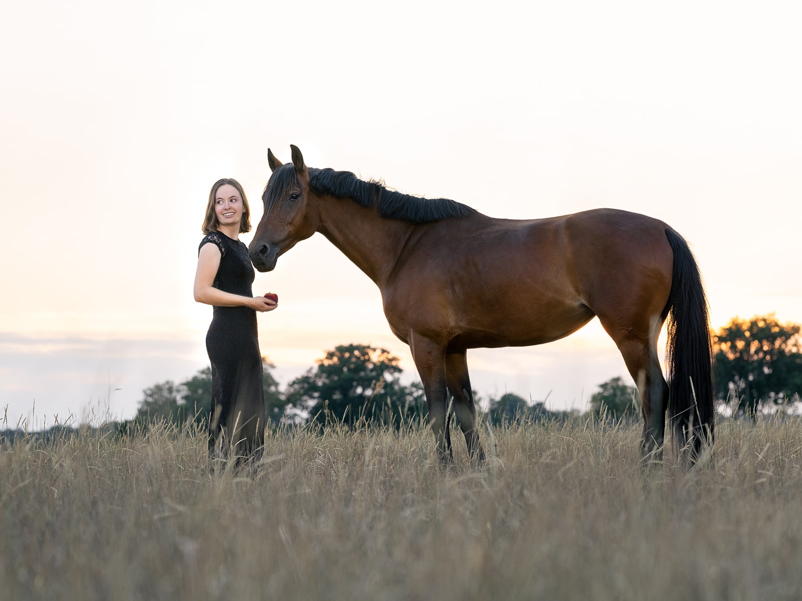 Woman in elegant black dress standing with her horse on evening pasture