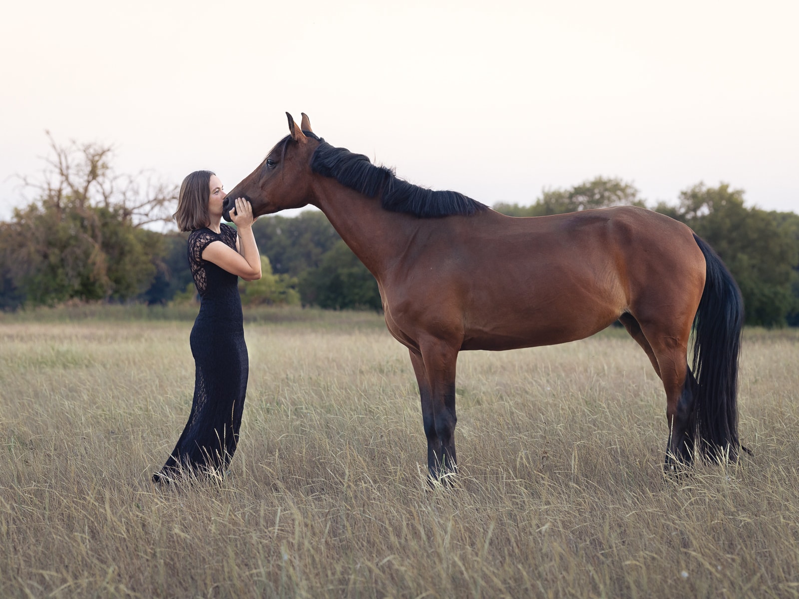 Woman in black lace dress kissing her brown horse on pasture