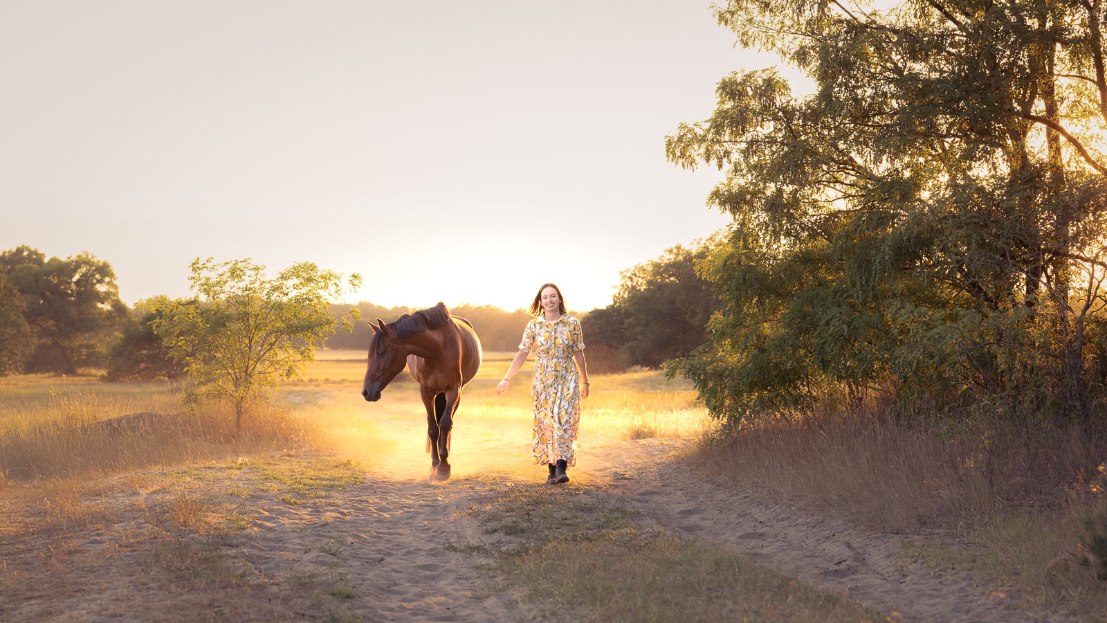 Woman in floral dress and brown horse walking together in golden sunset